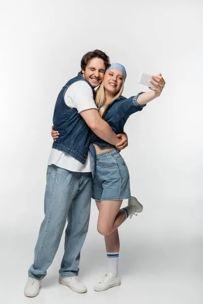 Cheerful man with closed eyes embracing woman taking selfie on smartphone on white — Stock Photo