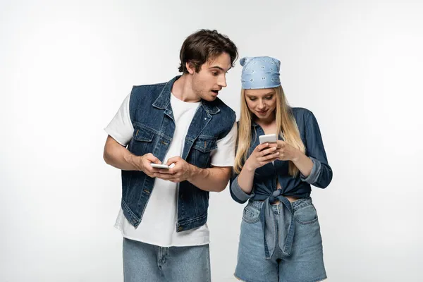 Здивований чоловік в джинсовому одязі, дивлячись на жінку, використовуючи мобільний телефон ізольований на білому — стокове фото
