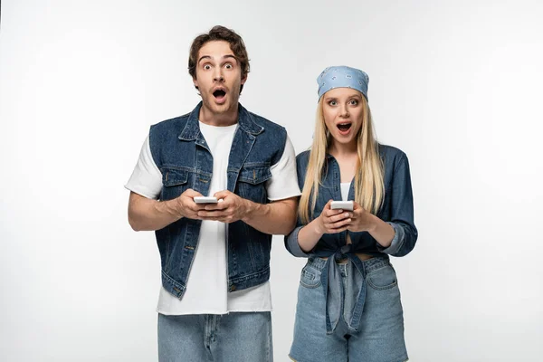 Erstauntes Paar in trendiger Jeanskleidung blickt in die Kamera, während es Mobiltelefone isoliert auf Weiß benutzt — Stockfoto