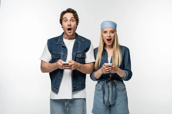 Casal surpreso em roupas de ganga na moda olhando para a câmera ao usar celulares isolados em branco — Fotografia de Stock