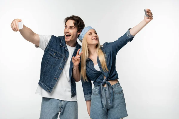 Excited man showing victory gesture while taking selfie on smartphones together with woman isolated on white — Stock Photo