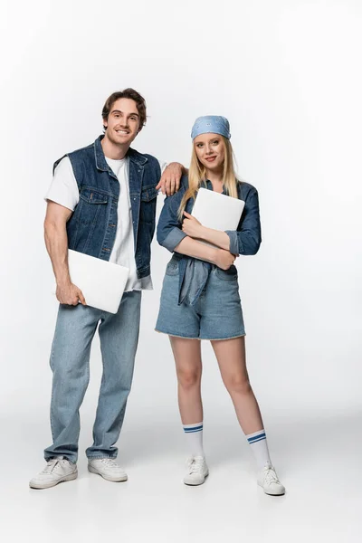 Par de freelancers felices en ropa vaquera sosteniendo computadoras portátiles en blanco - foto de stock