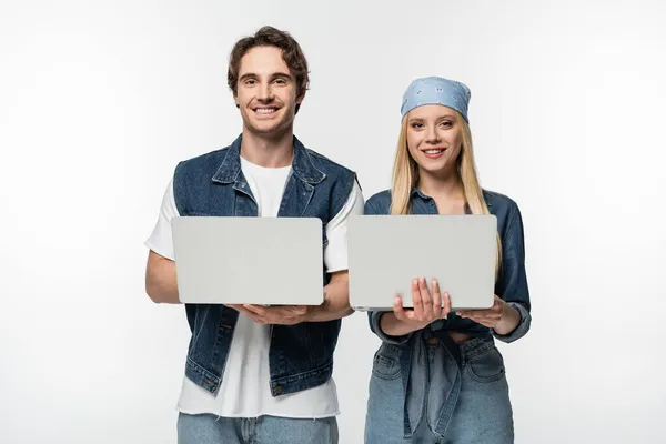 Coppia felice indossando vestiti di denim e tenendo i computer portatili isolati su bianco — Foto stock