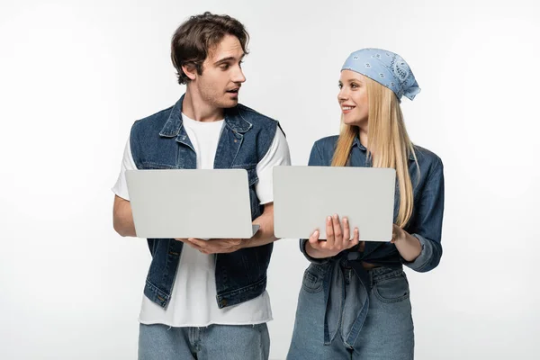 Casal feliz em roupas de ganga elegantes olhando uns para os outros ao usar laptops isolados em branco — Fotografia de Stock