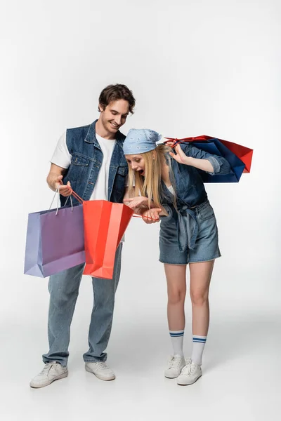 Mulher espantada olhando no saco de compras em mãos de namorado elegante no branco — Fotografia de Stock