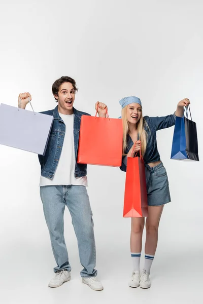 Felice e stupito coppia in denim abbigliamento che mostra multicolore shopping bags su bianco — Foto stock
