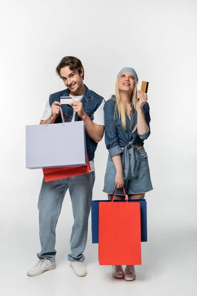 Couple élégant en vêtements en denim posant avec des sacs à provisions et des cartes de crédit sur blanc — Photo de stock