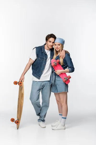 Casal na moda com longboard e penny board abraçando e olhando para a câmera no branco — Fotografia de Stock