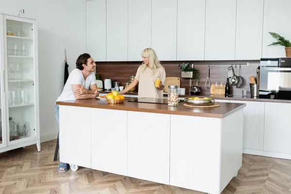 Happy man looking at cheerful girlfriend with glass of orange juice in kitchen — Stock Photo