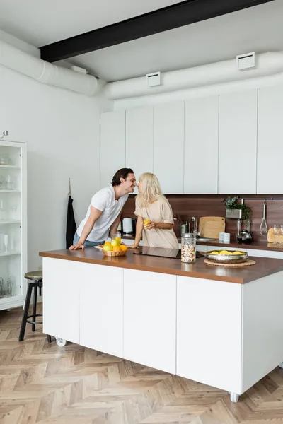 Feliz hombre besando alegre novia con vaso de jugo de naranja en cocina - foto de stock