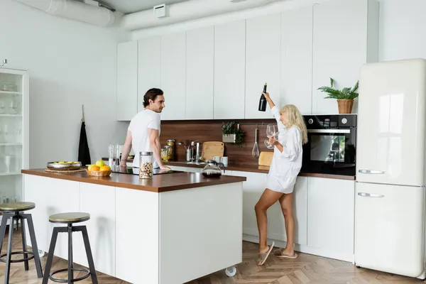 Fröhliche Frau hält Flasche Wein und Gläser neben Freund in Küche — Stockfoto