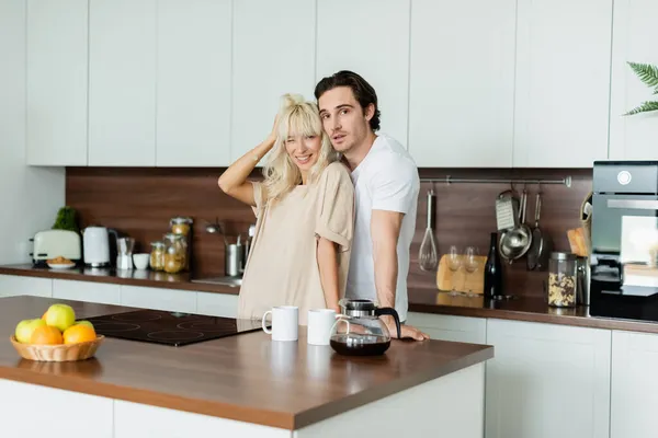 Mulher feliz ajustando cabelo loiro perto de namorado na cozinha — Fotografia de Stock