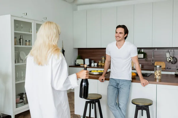 Homem feliz olhando para mulher loira com garrafa de vinho na cozinha — Fotografia de Stock