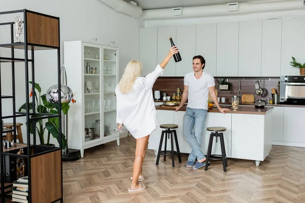 Uomo allegro guardando la donna bionda con bottiglia di vino e bicchieri in cucina — Foto stock