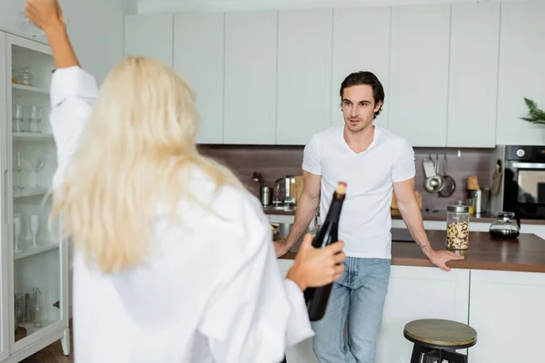 Tattooed man looking at blurred blonde woman with bottle of wine in kitchen — Stock Photo