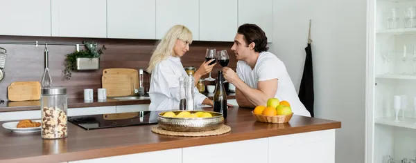 Pareja joven tintineo vasos de vino tinto en la cocina, pancarta - foto de stock