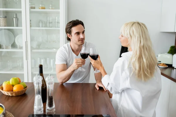 Joven hombre y mujer tintineo vasos de vino tinto en la cocina - foto de stock