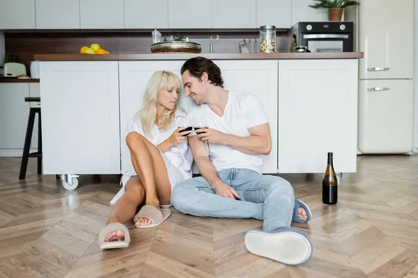 Homem feliz e mulher clinking copos de vinho tinto enquanto sentado no chão da cozinha — Fotografia de Stock