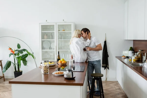 Jovem casal com copos de vinho abraçando na cozinha moderna — Fotografia de Stock
