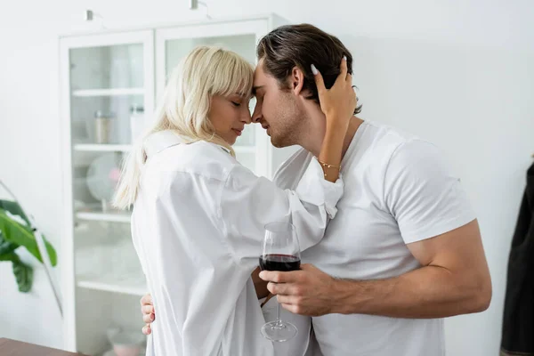 Vista lateral do jovem com copo de vinho abraçando com mulher loira na cozinha moderna — Fotografia de Stock