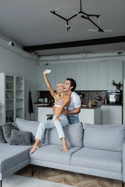 Mujer feliz tomando selfie con novio en sala de estar - foto de stock