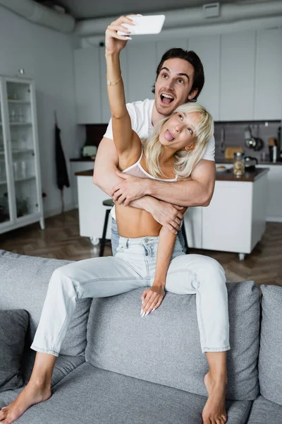 Donna bionda che sporge la lingua mentre prende selfie con il fidanzato felice in soggiorno — Foto stock