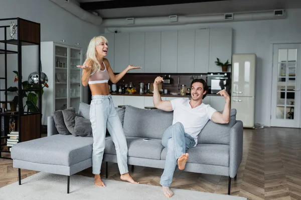 Happy woman gesturing near cheerful boyfriend pointing at himself while sitting on sofa — Stock Photo