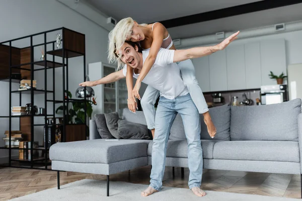 Homem feliz com as mãos estendidas piggybacking namorada loira na sala de estar — Fotografia de Stock