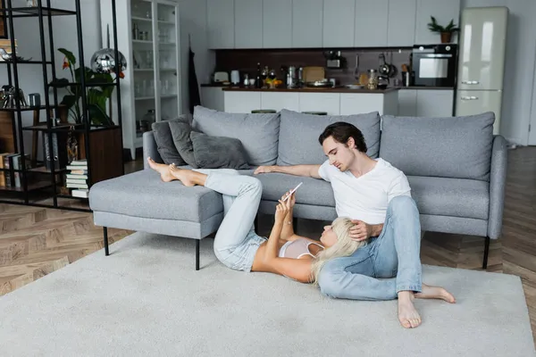 Mulher loira usando smartphone enquanto deitado perto do namorado na sala de estar — Fotografia de Stock