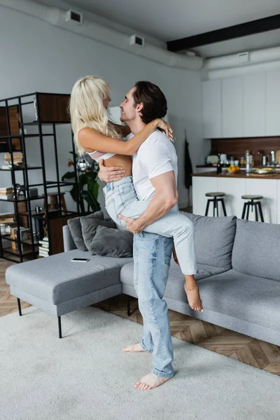 Homem feliz levantando namorada loira na sala de estar — Fotografia de Stock