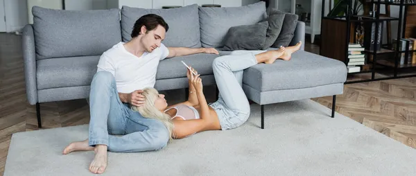 Blonde woman using smartphone while lying near boyfriend in living room, banner — Stock Photo