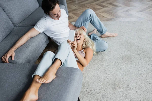 Cheerful woman laughing and using smartphone while lying near boyfriend in living room — Stock Photo