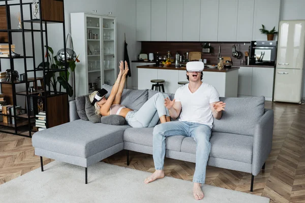 Blonde woman in vr headset lying on sofa with outstretched hands near astonished boyfriend — Stock Photo