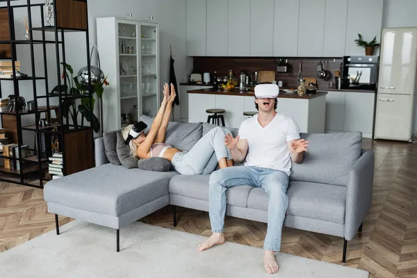 Femme blonde dans vr casque couché sur le canapé avec les mains tendues près de petit ami étonné — Photo de stock