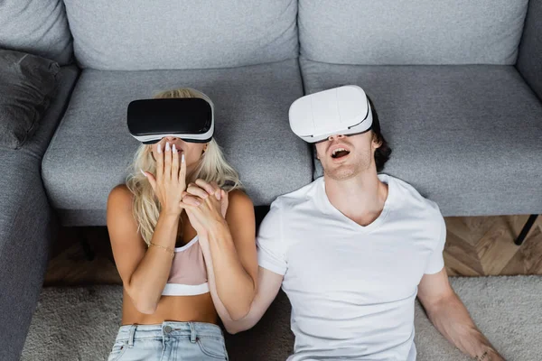 High angle view of shocked couple in vr headsets sitting near grey couch — Stock Photo