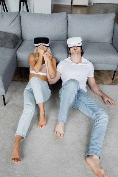 Shocked couple in vr headsets sitting on carpet near grey couch — Stock Photo