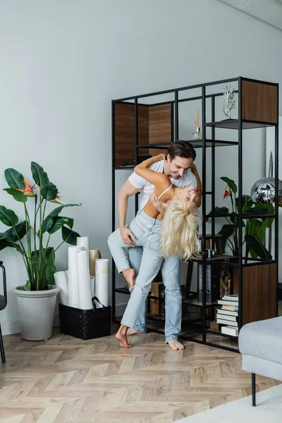 Comprimento total do homem feliz dobrando mulher alegre enquanto dança perto de rack na sala de estar — Fotografia de Stock