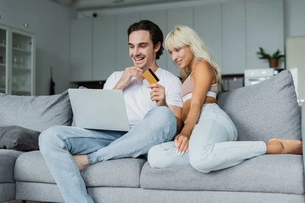 Hombre alegre celebración de la tarjeta de crédito cerca de la novia mientras mira el ordenador portátil y hacer compras en línea - foto de stock