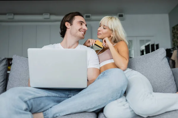 Casal feliz com cartão de crédito e laptop olhando uns para os outros fazendo compras on-line — Fotografia de Stock