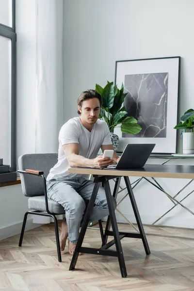 Jovem usando smartphone perto de laptop na mesa — Fotografia de Stock