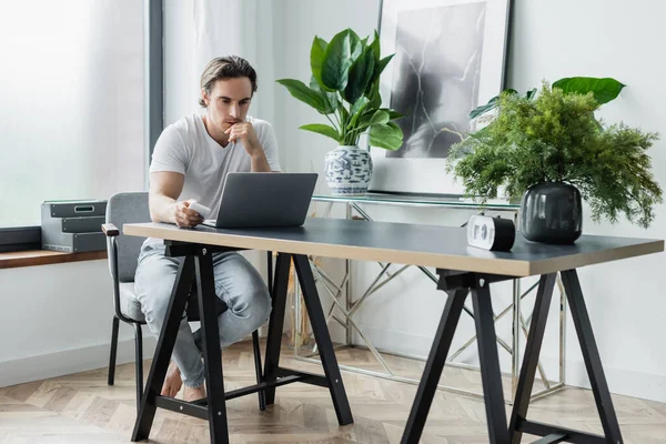 Concentrated freelancer looking at laptop while holding smartphone and working from home — Stock Photo
