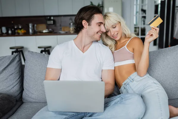 Mujer feliz celebración de la tarjeta de crédito cerca de novio alegre mientras que las compras en línea en casa - foto de stock