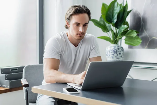 Konzentrierte Freiberuflerin schaut von zu Hause aus auf Laptop und Smartphone am Schreibtisch — Stockfoto