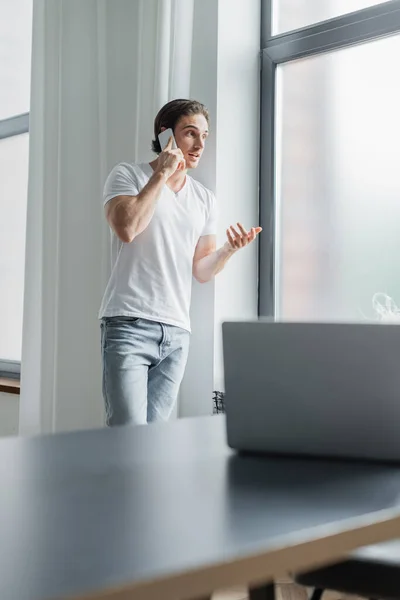Молодой человек жестикулирует во время разговора на смартфоне рядом с размытым ноутбуком на столе — стоковое фото