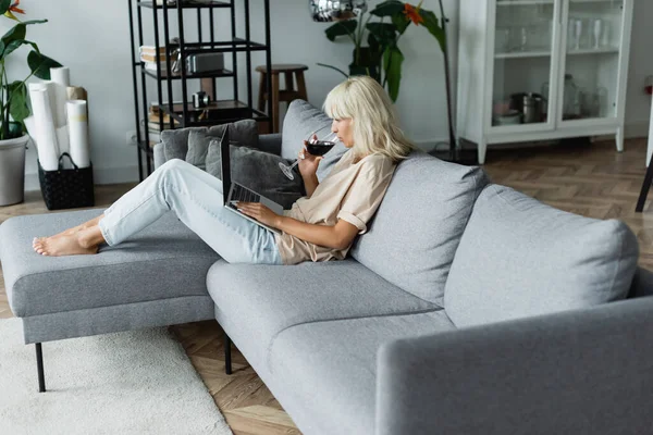 Blondine trinkt Rotwein, während sie Laptop im Wohnzimmer benutzt — Stockfoto