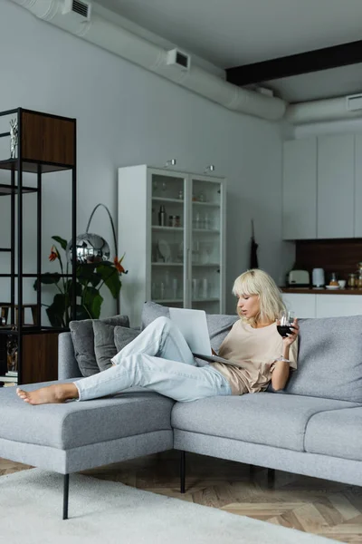 Blondine hält Glas Rotwein in der Hand, während sie Laptop im Wohnzimmer benutzt — Stockfoto