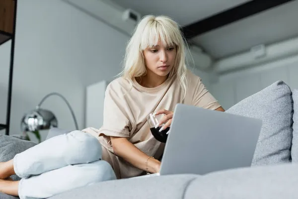 Blonde Freiberuflerin hält ein Glas Rotwein in der Hand, während sie Laptop im Wohnzimmer benutzt — Stockfoto