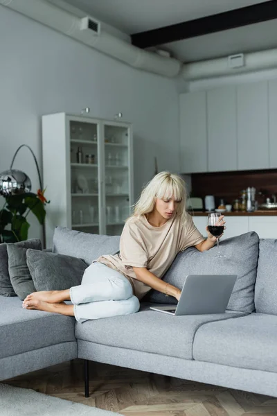 Barefoot at blonde woman holding glass of red wine while using laptop in living room — Stock Photo