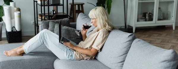 Femme blonde buvant du vin rouge tout en utilisant un ordinateur portable dans le salon, bannière — Photo de stock