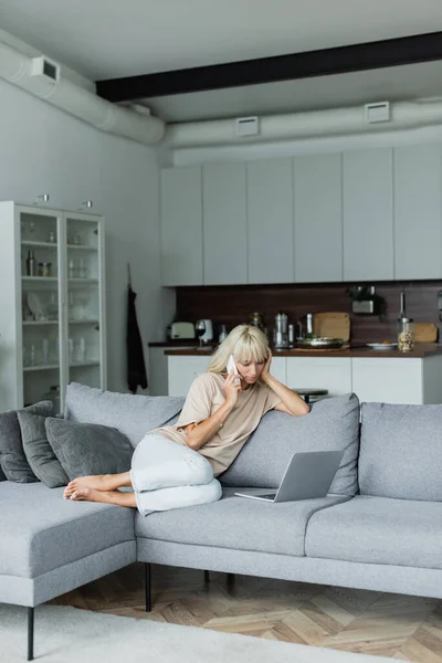 Barefoot and blonde freelancer talking on smartphone near laptop on couch — Stock Photo